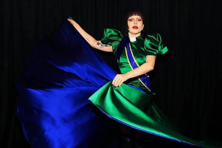Uma modelo está posando em um fundo preto, vestindo um vestido de gala com mangas volumosas. O vestido é predominantemente verde com detalhes em azul, e a modelo está segurando uma parte do vestido, fazendo com que ele se espalhe. Ela tem cabelo curto e usa maquiagem marcante, com lábios vermelhos.