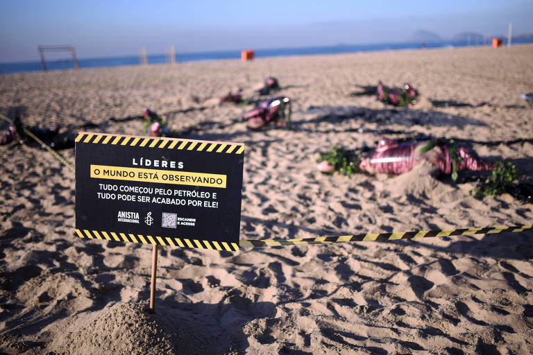 A imagem mostra uma praia com areia clara e um mar ao fundo. No primeiro plano, há um cartaz preto e amarelo com a inscrição "LÍDERES O MUNDO ESTÁ OBSERVANDO TUDO COMEÇOU PELO PETRÓLEO E TUDO PODE SER ACABADO POR ELE!". O cartaz está cercado por uma fita de segurança. Ao fundo, são visíveis alguns objetos cobertos com flores, sugerindo uma intervenção artística ou uma instalação. O céu está limpo e a cena transmite uma sensação de tranquilidade, contrastando com a mensagem do cartaz.