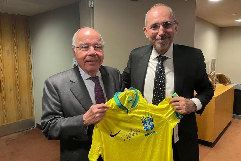 Duas pessoas estão em um ambiente interno, segurando uma camisa amarela da Seleção Brasileira de Futebol. A camisa possui o escudo da seleção e uma assinatura. A pessoa à esquerda está vestindo um terno escuro e uma gravata, enquanto a pessoa à direita está usando um terno claro e uma gravata preta com pontos brancos. Ao fundo, há uma parede e uma mesa.