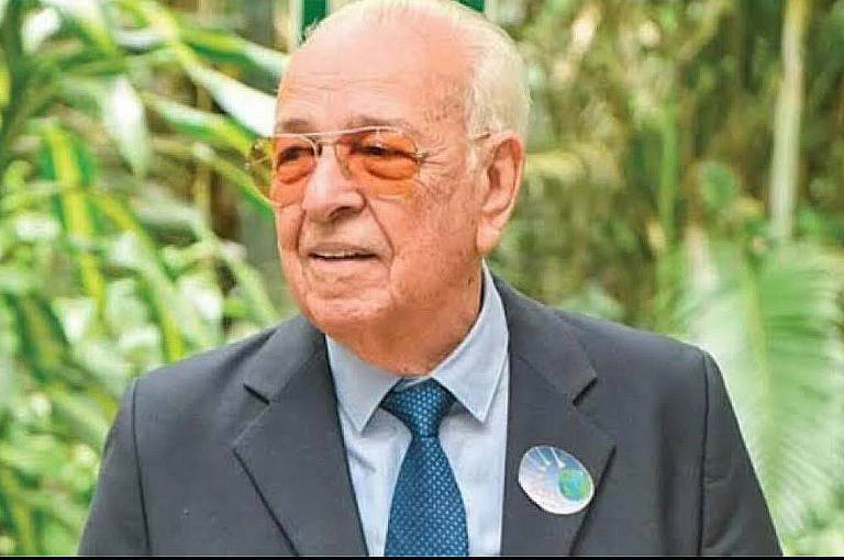 Um homem idoso está posando em um ambiente natural, cercado por plantas verdes. Ele usa um terno escuro, uma camisa clara e óculos de sol laranja. Na lapela do seu paletó, há um botão com um desenho que parece representar o planeta Terra.