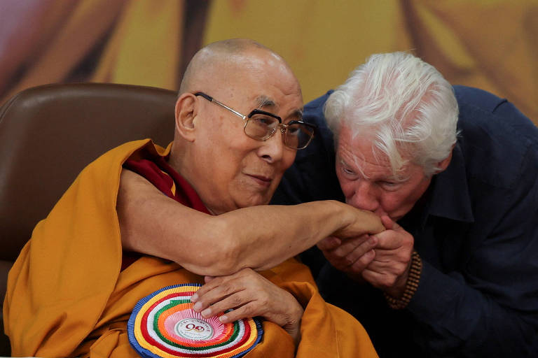 A imagem mostra o Dalai Lama sentado em uma cadeira, sorrindo e olhando para um homem que está se inclinando para beijar seu braço. O Dalai Lama está vestido com um manto laranja e vermelho, e usa óculos. O homem tem cabelo grisalho e está vestido com uma camisa escura. Ao fundo, há uma imagem desfocada que sugere um ambiente de evento ou conferência.