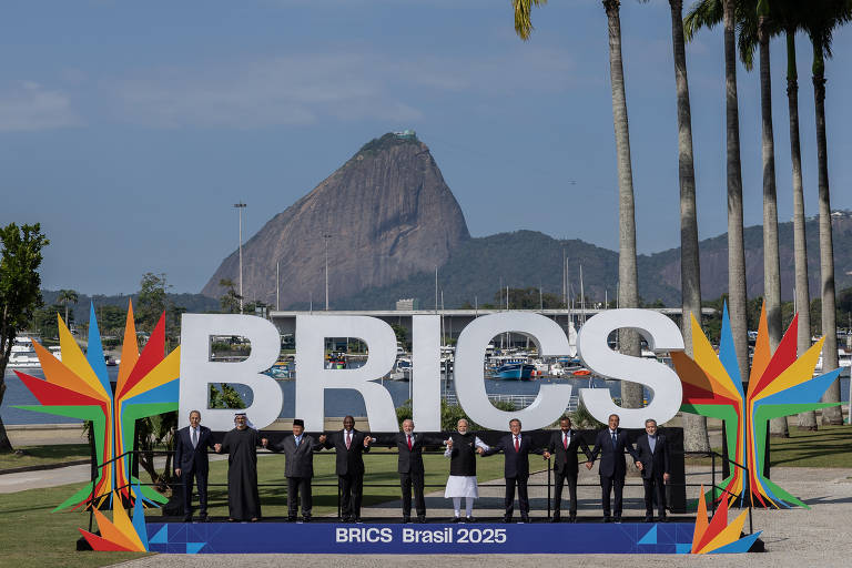 A imagem mostra um grupo de pessoas em pé em frente a uma grande estrutura com a palavra 'BRICS' em destaque. Ao fundo, há uma montanha e um corpo d'água. As pessoas estão vestidas formalmente e algumas seguram bandeiras. A estrutura é colorida, com elementos gráficos em vermelho, amarelo e verde, e há palmeiras ao redor.