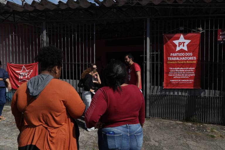A imagem mostra um grupo de pessoas em frente a um prédio com uma grade. Há uma bandeira do Partido dos Trabalhadores (PT) visível na parede, com um texto em destaque. Algumas pessoas estão conversando e outras observando. O ambiente parece ser um local de encontro ou evento político.
