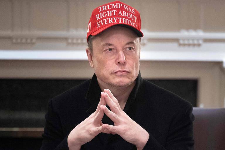 Um homem está sentado à mesa com as mãos entrelaçadas. Ele usa um boné vermelho com a frase "TRUMP WAS RIGHT ABOUT EVERYTHING!". O ambiente parece ser um escritório ou sala de reuniões, com uma parede clara ao fundo e uma lareira visível.