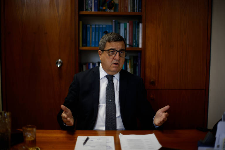 Um homem de terno e gravata está sentado à mesa, gesticulando com as mãos enquanto fala. Ele usa óculos e tem cabelo curto e grisalho. Ao fundo, há uma estante com livros e documentos. Na mesa, há uma folha de papel e um copo com bebida.