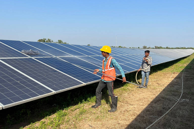 Dois trabalhadores estão em um campo de painéis solares. Um deles, usando um capacete amarelo e colete laranja, está puxando um cabo, enquanto o outro, vestido com uma camisa xadrez, observa. O céu está claro e há vegetação ao redor dos painéis.