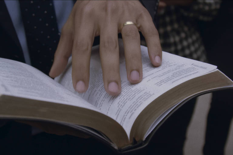 A imagem mostra uma mão masculina segurando uma Bíblia aberta. A mão está posicionada sobre as páginas do livro, e o homem está vestido com um terno escuro. Ao fundo, é possível ver outras pessoas, mas não estão claramente focadas. A iluminação é suave, destacando a Bíblia e a mão que a segura.