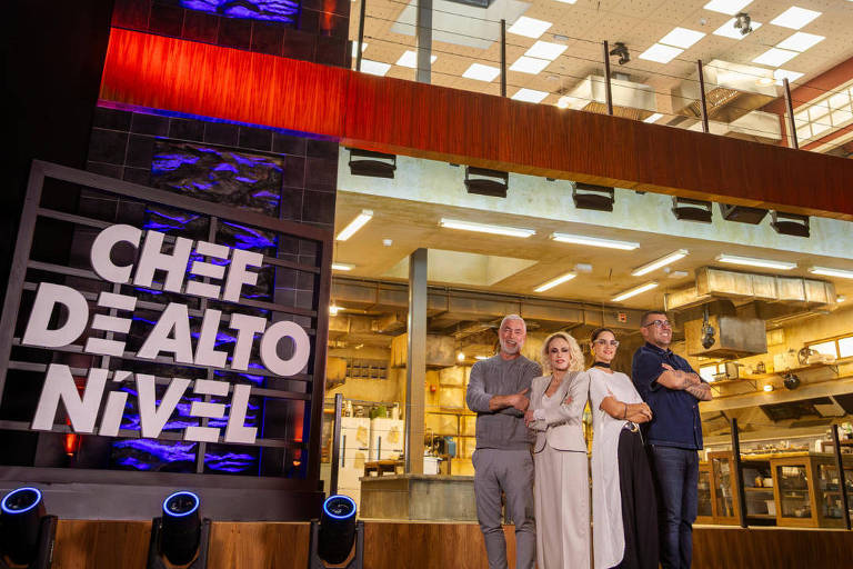 A imagem mostra quatro pessoas em frente a um grande letreiro que diz 'CHEF DE ALTO NÍVEL'. Ao fundo, há uma estrutura moderna com janelas amplas e iluminação interna visível. As pessoas estão posando para a foto, com expressões sorridentes, e vestem roupas casuais. O ambiente parece ser um estúdio ou um espaço de evento relacionado à gastronomia.