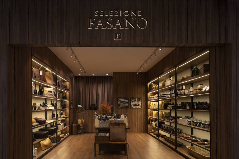 A imagem mostra a entrada de uma loja chamada 'Selezione Fasano'. A fachada é feita de madeira escura e possui uma iluminação suave. Dentro da loja, há prateleiras organizadas com diversos produtos, incluindo bolsas, garrafas e itens de decoração. No centro, há uma mesa com algumas mercadorias expostas. O ambiente é elegante e acolhedor.