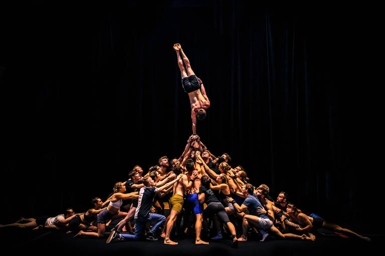 Um grupo de acrobatas está em uma formação complexa, com várias pessoas em diferentes posições. No centro, um acrobata está sendo levantado por outros, enquanto muitos outros estão em volta, apoiando-se uns nos outros. A cena é iluminada dramaticamente, com um fundo escuro que destaca a performance. As pessoas estão vestidas com roupas de ginástica coloridas, e a composição transmite uma sensação de força e união.