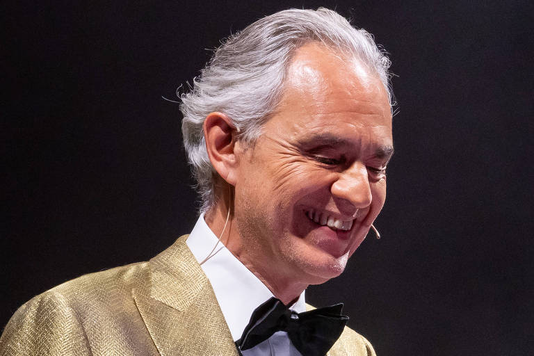 Um homem sorridente está vestido com um paletó dourado e uma gravata borboleta preta. Ele tem cabelo grisalho e está em um ambiente com fundo escuro, sugerindo um evento formal ou apresentação. O homem parece estar em um momento de descontração, possivelmente interagindo com o público.