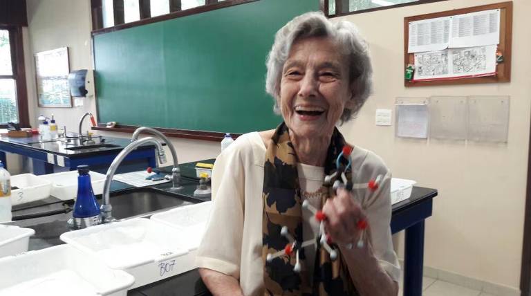 A imagem mostra uma mulher idosa sorrindo em um laboratório. Ela está segurando um modelo molecular colorido, com esferas vermelhas, brancas e azuis. Ao fundo, há uma lousa verde e mesas de laboratório com equipamentos e recipientes. A mulher usa uma blusa clara e um lenço estampado.