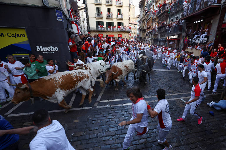 A imagem mostra uma cena da famosa corrida de touros em Pamplona, na Espanha. Várias pessoas vestidas de branco com lenços vermelhos estão correndo pelas ruas, enquanto touros e vacas estão sendo conduzidos. O ambiente é festivo, com uma multidão assistindo das varandas e do lado da rua. O chão é de paralelepípedos e as construções ao fundo são típicas da arquitetura espanhola.