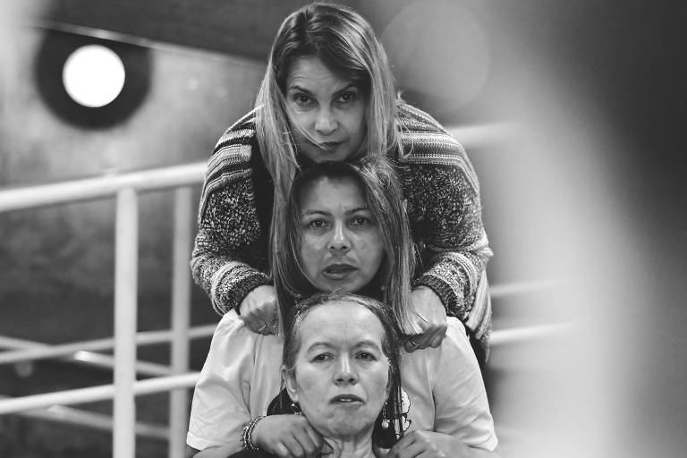 A imagem em preto e branco mostra três mulheres em uma formação vertical. A mulher na parte superior está com as mãos nos ombros da mulher do meio, que, por sua vez, está olhando para a câmera. A mulher na parte inferior está com uma expressão neutra e usa uma camiseta com a frase 'self'. O fundo é desfocado, com uma estrutura que parece ser uma escada e uma luz circular visível.
