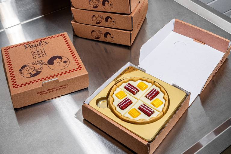 A imagem mostra uma caixa de pizza aberta, revelando uma pizza de sobremesa decorada com fatias de frutas coloridas. A caixa é de papelão marrom com uma tampa que possui um design simples. Ao fundo, há várias caixas empilhadas, também de papelão marrom, em uma superfície metálica. A pizza parece ser feita de um material que imita a aparência de uma pizza real, mas é claramente uma sobremesa.