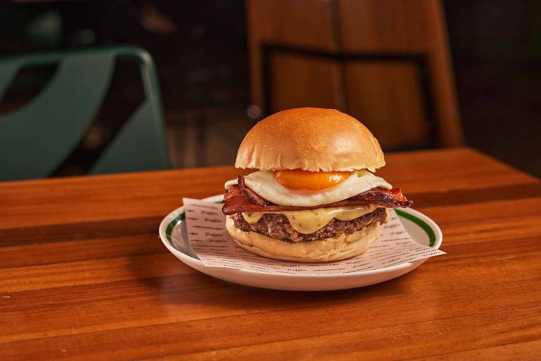 Um hambúrguer servido em um prato branco, com um pão macio, carne, queijo derretido, bacon e um ovo frito em cima. O fundo é uma mesa de madeira e há uma cadeira verde ao fundo.