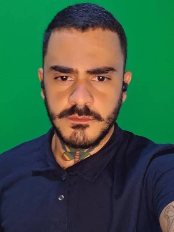 Um homem está posando para a foto, com um fundo verde. Ele tem cabelo curto e uma barba bem cuidada. O homem usa uma camiseta polo de cor escura e possui tatuagens visíveis nos braços e no pescoço. Ele está olhando diretamente para a câmera com uma expressão séria.