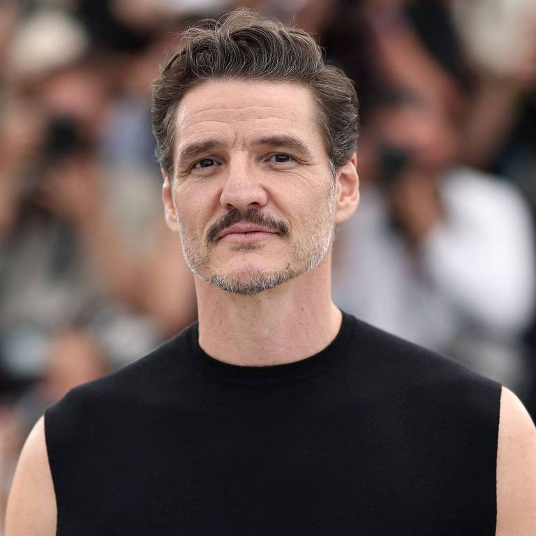 Um homem com cabelo castanho escuro e barba, vestindo uma camiseta sem mangas preta, está posando para a câmera. Ao fundo, há várias pessoas com câmeras, sugerindo que ele está em um evento público ou festival. A expressão do homem é séria e confiante.