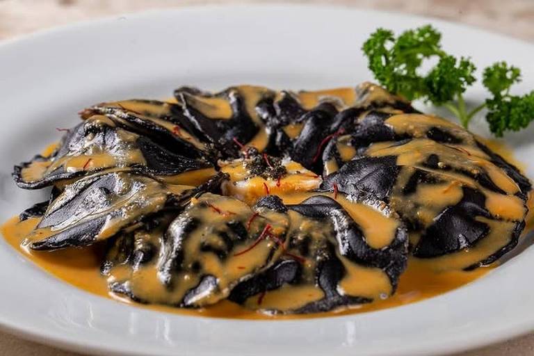 Um prato de comida gourmet apresenta uma porção de massa escura, possivelmente ravioli, coberta com um molho cremoso de cor amarela. O prato é decorado com um raminho de salsa verde ao lado. A apresentação é sofisticada, com a massa disposta de forma circular e o molho espalhado de maneira uniforme.