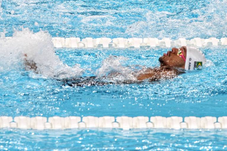 Um nadador está competindo em uma piscina, com água azul clara ao fundo. Ele está de costas, usando um boné de natação branco e óculos. A água está espirrando ao redor dele enquanto ele se move.