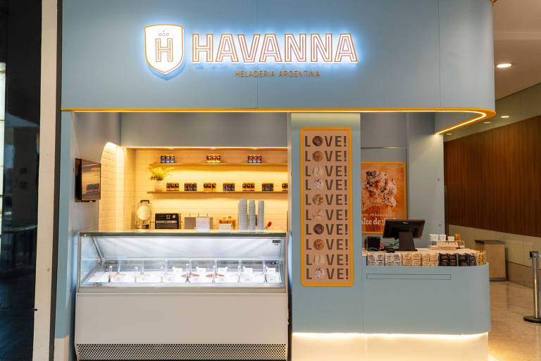 A imagem mostra a fachada de uma loja da Havanna, com um letreiro iluminado em azul e branco. A loja possui uma vitrine com produtos expostos, incluindo doces e chocolates. Ao lado da vitrine, há uma parede com uma mensagem que diz 'LOVE!' repetida em letras grandes. O ambiente é moderno e bem iluminado, com um balcão de atendimento visível.
