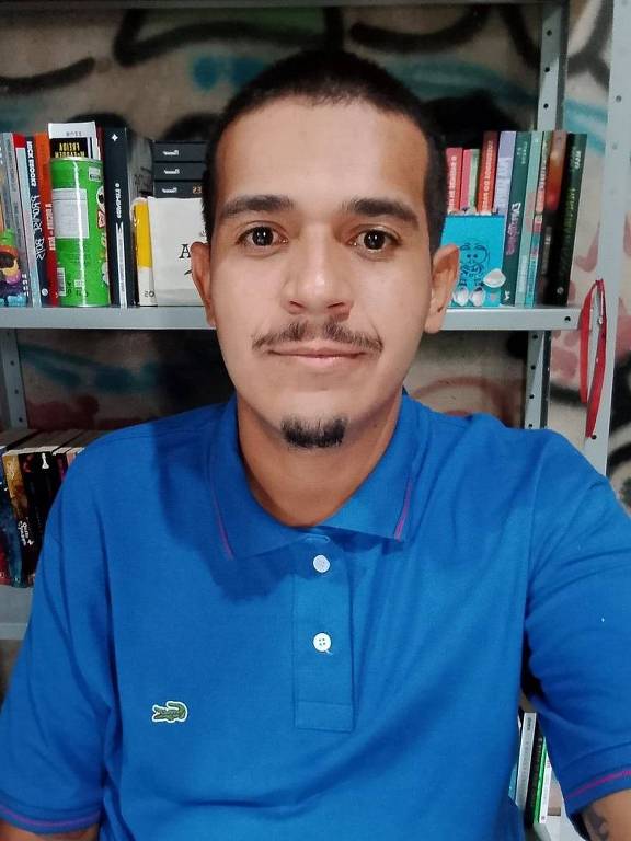 Um homem está posando para a foto em um ambiente interno. Ele usa uma camisa polo azul e tem um leve sorriso. Ao fundo, há uma estante com livros e outros objetos. A parede atrás dele tem grafites ou pinturas.