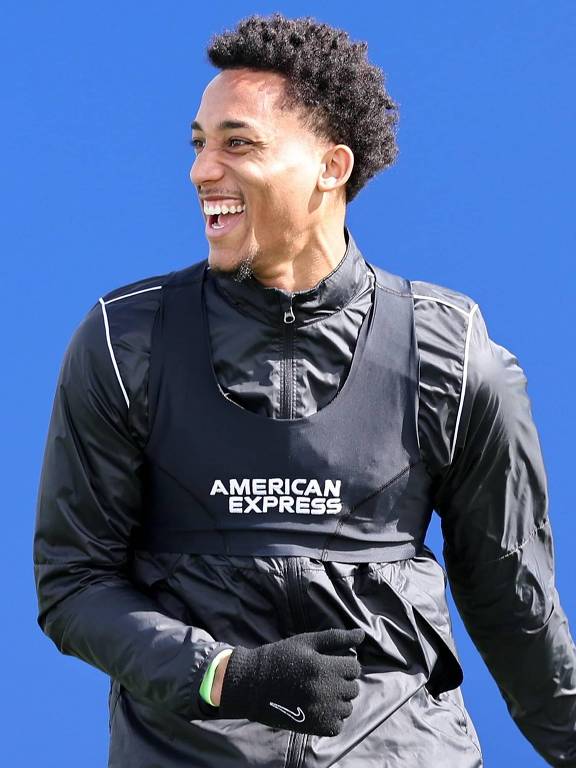 Um jogador de futebol está sorrindo enquanto treina. Ele usa uma camisa preta com um colete que tem a inscrição 'AMERICAN EXPRESS'. O fundo é azul, e o jogador tem cabelo crespo e curto. Ele também está usando uma luva na mão esquerda.