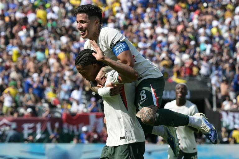 Na imagem, dois jogadores de futebol estão comemorando um gol. Um jogador está em pé, com as mãos levantadas, enquanto o outro está agachado, sendo carregado nas costas. Ao fundo, há uma multidão de torcedores em um estádio, com bandeiras e camisas de times. A atmosfera é de celebração e alegria.