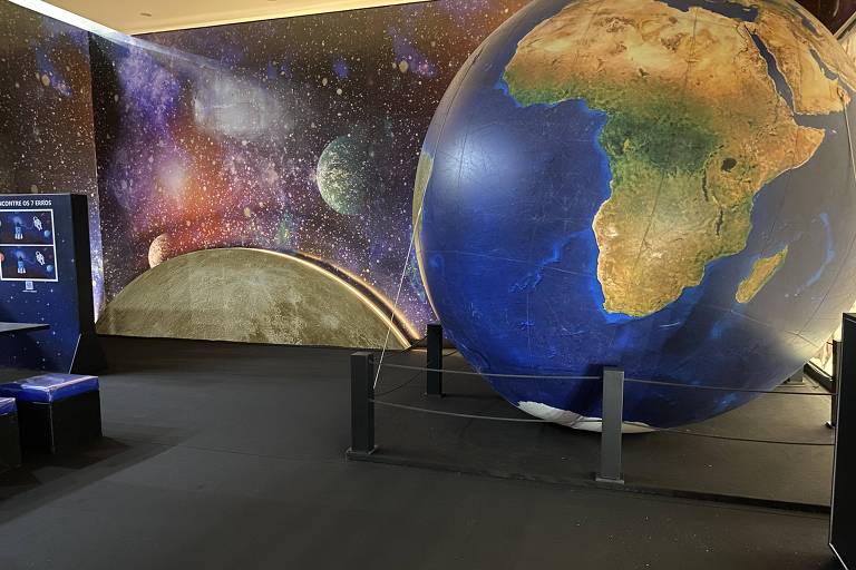 A imagem mostra uma sala de exposição com um grande globo terrestre em destaque, representando a África. O fundo da sala é decorado com um mural espacial que inclui planetas e estrelas. Há também algumas mesas e bancos azuis dispostos no espaço.