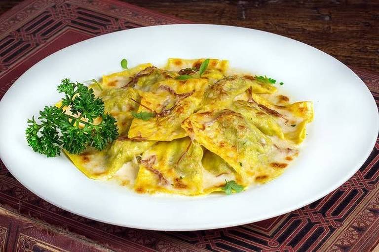 Um prato de ravioli servido em um prato branco. Os raviolis são cobertos com um molho cremoso e estão dispostos de forma organizada. Há uma guarnição de salsa ao lado dos raviolis. O fundo do prato é decorado com um padrão em tons de vermelho e dourado.