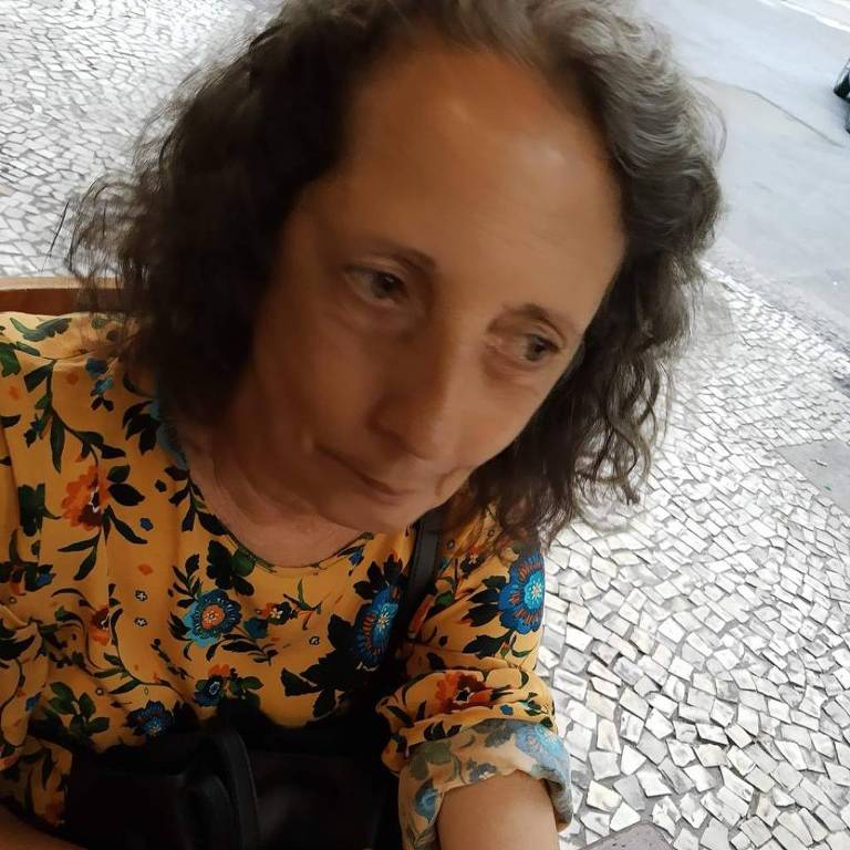 A imagem mostra uma pessoa com cabelo cacheado e uma blusa colorida, sentada em uma mesa. A pessoa está olhando para a câmera e tem uma tatuagem em forma de quadrado visível em seu braço. O fundo apresenta um piso de pedras e um ambiente externo.