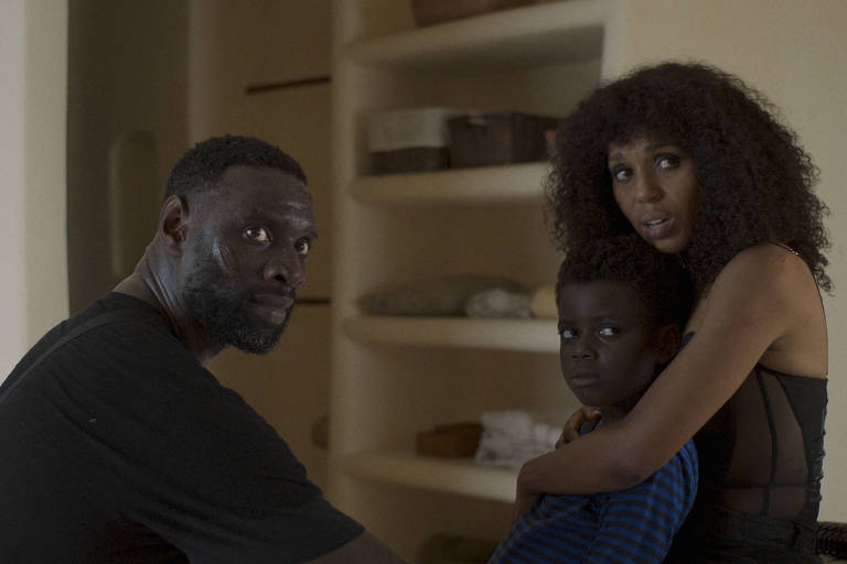 A imagem mostra um homem e uma mulher sentados em um ambiente interno, com prateleiras ao fundo. O homem, de pele escura e cabelo curto, está olhando para a câmera com uma expressão séria. A mulher, com cabelo cacheado e vestido preto, está segurando uma criança, que parece estar assustada. A criança, de pele escura, está vestindo uma camiseta listrada azul e preta. O ambiente é iluminado de forma suave, criando uma atmosfera tensa.