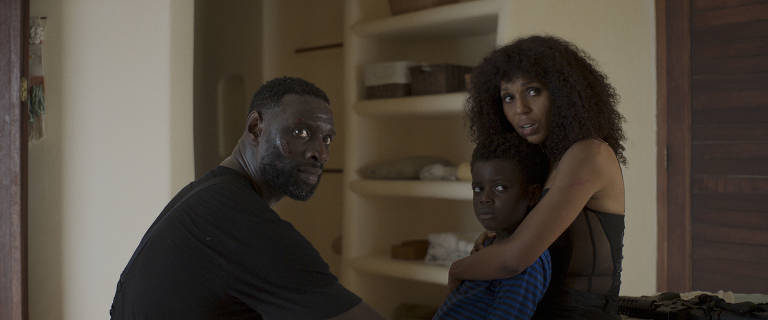 A imagem mostra um homem e uma mulher sentados em um ambiente interno, com prateleiras ao fundo. O homem, de pele escura e cabelo curto, está olhando para a câmera com uma expressão séria. A mulher, com cabelo cacheado e vestido preto, está segurando uma criança, que parece estar assustada. A criança, de pele escura, está vestindo uma camiseta listrada azul e preta. O ambiente é iluminado de forma suave, criando uma atmosfera tensa.
