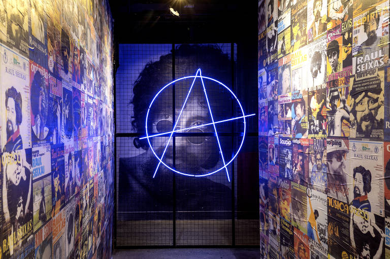 A imagem mostra um corredor decorado com pôsteres de artistas, predominantemente de Raúl Seixas, nas paredes. No fundo, há uma grande imagem em preto e branco de uma pessoa com óculos. No centro do corredor, uma luz neon azul em forma de símbolo anarquista destaca-se contra a grade que separa o espaço.