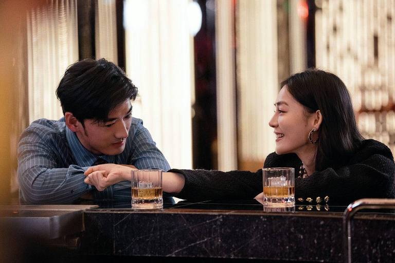 The image shows a couple sitting at a bar. The man, with dark hair and a blue shirt, is looking at the woman, who has long, dark hair and a black blouse. They are touching, their hands intertwined, as they talk. On the bar counter, there are two glasses of drinks. The room is lit with bright lights in the background, creating a cozy atmosphere.