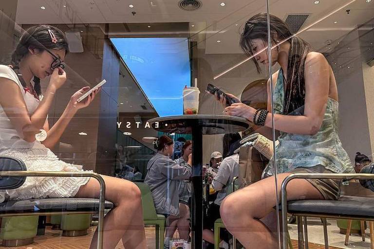 The image shows two women sitting in a cafe, each using a cell phone. They are seated at a glass table, with a brightly lit room in the background. The woman on the left is wearing a white blouse and skirt, while the woman on the right is wearing a light blue blouse and shorts. The setting appears modern, with other people visible in the background.