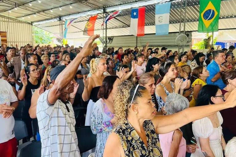 A imagem mostra uma grande multidão em um evento religioso, com pessoas de diversas idades e etnias. Muitas delas estão de pé, com as mãos levantadas, em um ambiente coberto. Ao fundo, há bandeiras de diferentes países penduradas, incluindo a bandeira do Brasil. A atmosfera parece ser de celebração ou adoração.