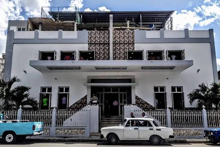 A imagem mostra a fachada de um edifício moderno, com uma estrutura simétrica e janelas retangulares. O prédio é de cor branca com detalhes em cinza e possui um padrão decorativo na parte superior. Há plantas na frente e alguns carros estacionados, incluindo um carro azul e um carro branco. O céu está claro com algumas nuvens.