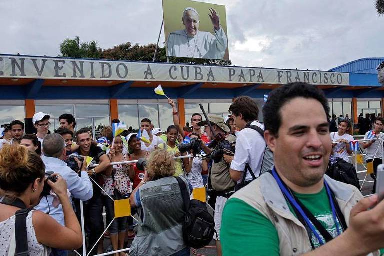A imagem mostra uma multidão reunida em um local de recepção, com um grande banner que diz 'Bienvenido a Cuba Papa Francisco'. Acima do banner, há uma imagem do Papa Francisco. As pessoas na multidão estão tirando fotos e algumas estão segurando celulares. O clima parece nublado e há uma atmosfera de expectativa e celebração.