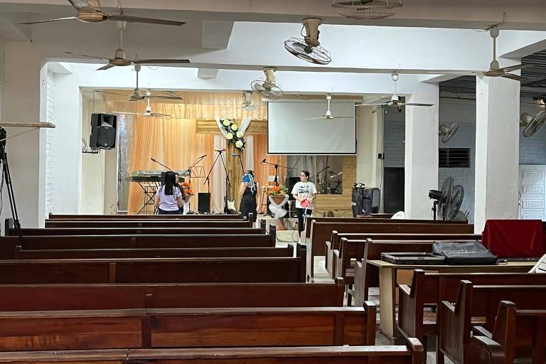 A imagem mostra o interior de uma igreja com bancos de madeira dispostos em fileiras. No fundo, há um altar com uma tela e um arranjo de flores. Algumas pessoas estão sentadas em um dos bancos, enquanto outras estão em pé perto do altar. Equipamentos de som e câmeras estão visíveis ao longo das paredes. O ambiente é bem iluminado, com ventiladores de teto visíveis.