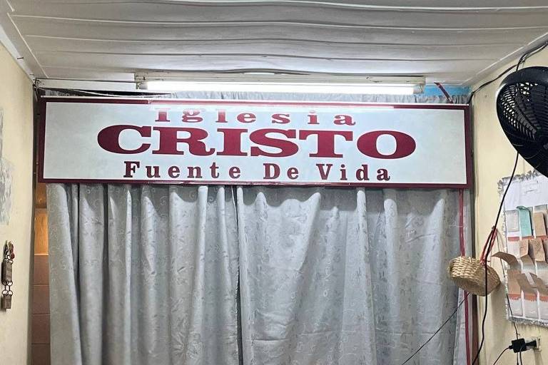 A imagem mostra o interior de um espaço com uma cortina clara e um ventilador de parede. Acima da cortina, há uma placa com o texto 'Iglesia CRISTO Fuente De Vida' em letras grandes e vermelhas sobre um fundo branco. O ambiente parece ser um local de culto ou reunião.