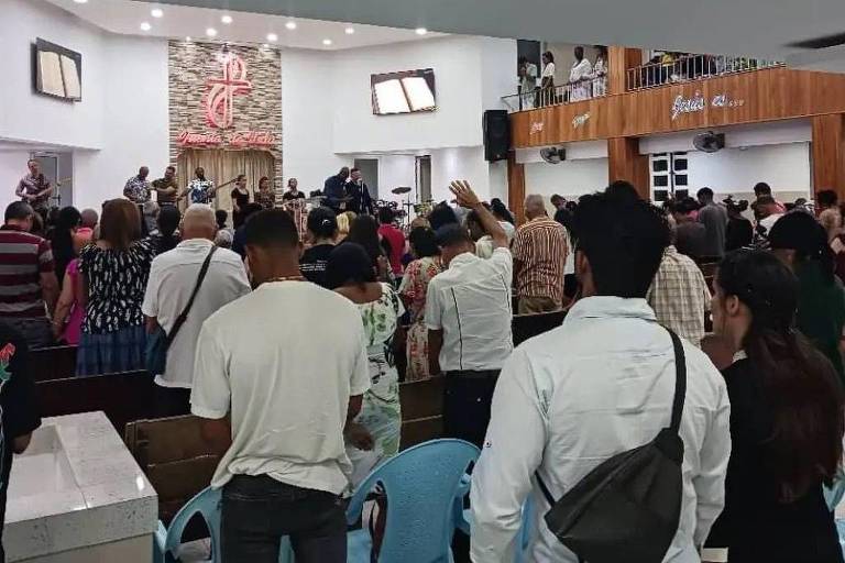 A imagem mostra um culto em uma igreja, com um grande número de pessoas em pé, algumas levantando as mãos. No fundo, há uma banda tocando instrumentos musicais. O ambiente é iluminado e decorado com um painel de madeira e uma tela. As pessoas estão vestidas de maneira casual e variada, e há um clima de celebração e adoração.