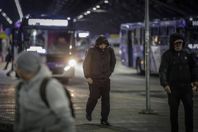 A imagem mostra um terminal de ônibus à noite, com várias pessoas caminhando. Um homem vestido de preto, com capuz, está em destaque, enquanto outros passageiros se movem ao fundo. Vários ônibus estão estacionados, e as luzes do terminal iluminam a cena, criando um ambiente urbano e noturno.