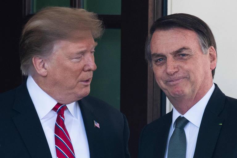 A imagem mostra dois homens em um ambiente interno, aparentemente em uma reunião. Um deles está usando um terno escuro com uma gravata listrada em vermelho e branco, enquanto o outro está vestido com um terno escuro e uma camisa branca. Ambos estão sorrindo e olhando um para o outro, com uma porta ao fundo