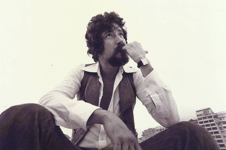 A imagem mostra um homem sentado, com cabelo encaracolado e barba, olhando para o lado enquanto leva a mão à boca em um gesto pensativo. Ele usa uma camisa branca e um colete escuro, e está em um ambiente ao ar livre, com prédios ao fundo. A foto é em preto e branco, o que dá um ar nostálgico à cena.