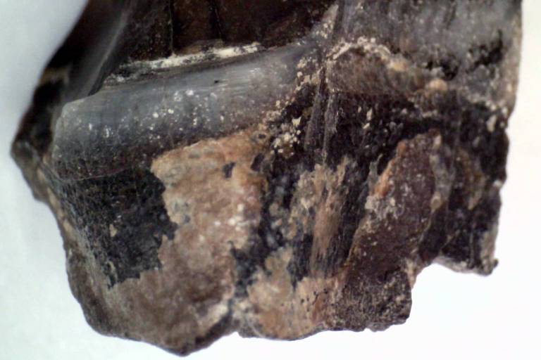 A imagem mostra um pedaço de obsidiana, uma rocha ígnea vítrea, com uma superfície brilhante e irregular. A rocha apresenta uma coloração predominantemente escura, com variações de tons que vão do preto ao marrom. A textura é lisa em algumas áreas e áspera em outras, evidenciando as características naturais da obsidiana.