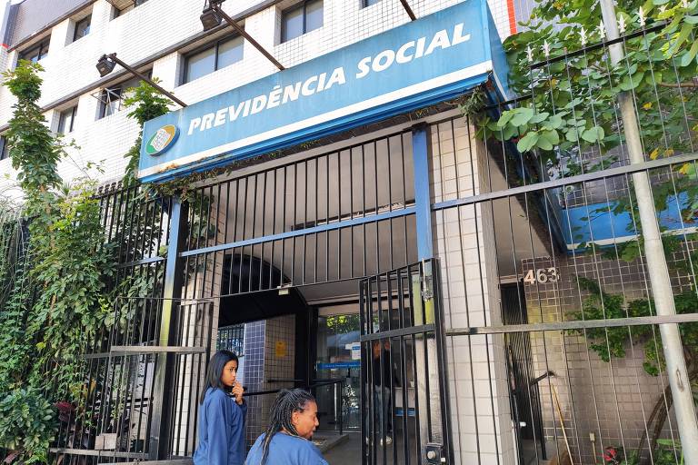 A imagem mostra a entrada de um prédio da Previdência Social, com uma placa azul na parte superior que diz 'PREVIDÊNCIA SOCIAL'. Há duas pessoas em frente ao portão, que está fechado, e o ambiente é cercado por uma grade. O local é cercado por vegetação, com algumas plantas visíveis ao redor da entrada.