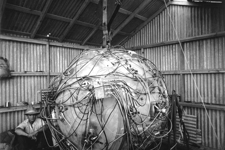 A imagem mostra uma esfera grande, coberta de fios e cabos, dentro de um galpão com paredes de metal ondulado. Um homem está sentado ao lado da esfera, observando o equipamento. O ambiente parece ser um local de testes ou experimentos científicos, com iluminação natural entrando pelo telhado.