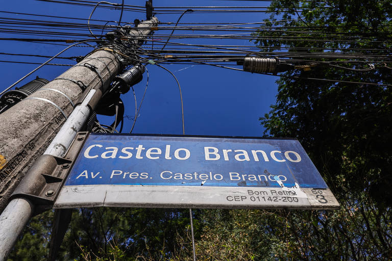 A imagem mostra uma placa de sinalização de rua com o nome "Castelo Branco" e a informação "Av. Pres. Castelo Branco". A placa está fixada em um poste de luz, que possui fios elétricos visíveis. O céu está claro e azul ao fundo, e há vegetação ao redor.
