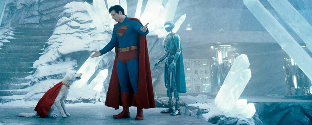 Cena do filme  Superman com David Corenswet  *** Local Caption *** A imagem mostra um homem vestido como Superman, com um traje azul e uma capa vermelha, interagindo com um cão que também está usando uma capa vermelha. O cenário é uma caverna de gelo com grandes cristais de gelo ao fundo. Ao lado do Superman, há um robô de cor azul. O ambiente é iluminado de forma suave, destacando os cristais e a neve ao redor.